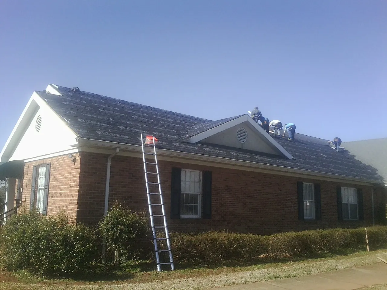 Crew tearing shingles off a brick ranch roof with a ladder set — Emergency Roof Repair work by our crew in Mc Farland