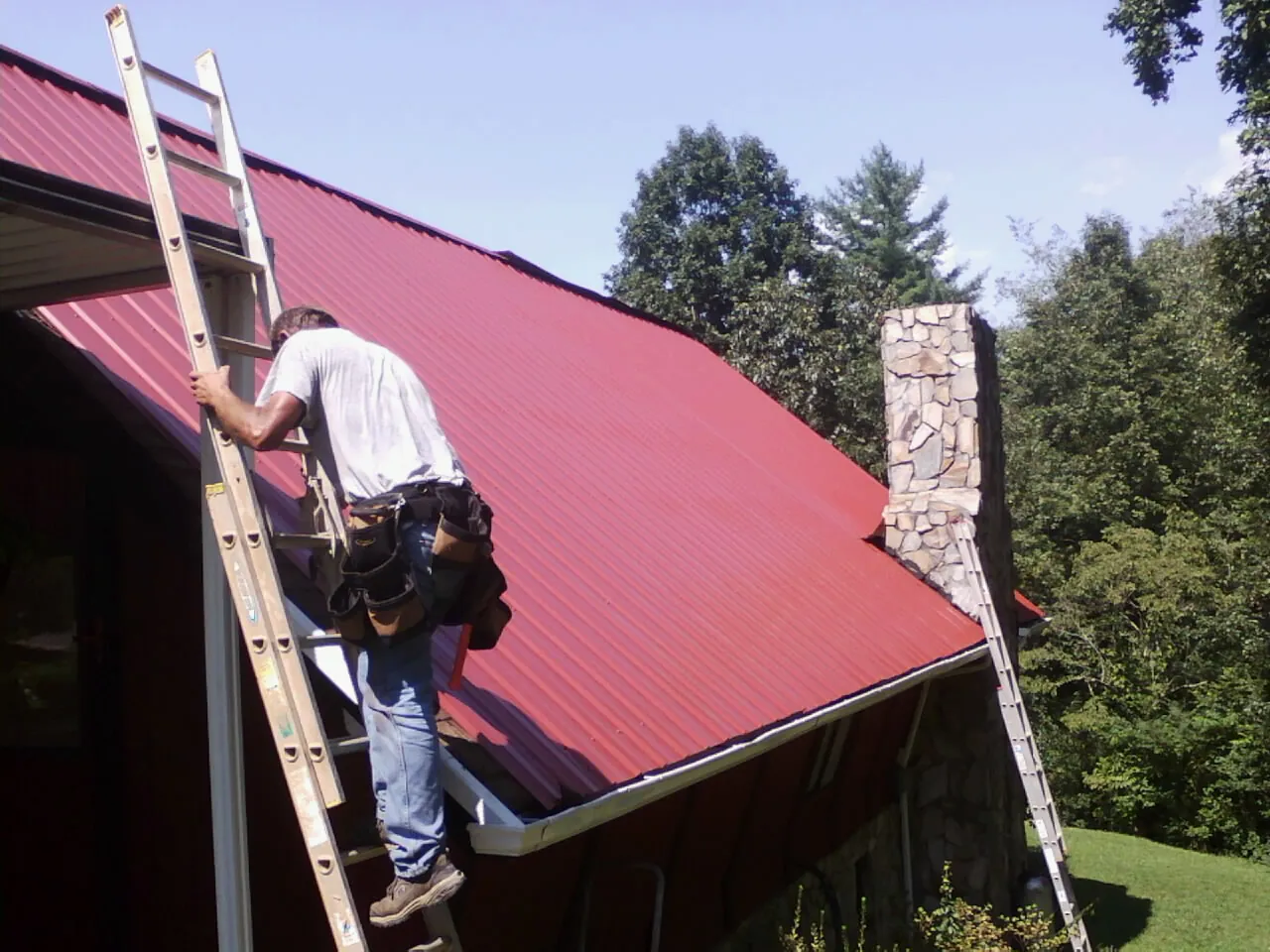 Roofer on a ladder beside a red metal roof and stone chimney — Soffit & Fascia Repair by Roofing Brothers in Mc Farland